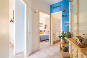 Un couloir mène à une chambre à coucher qui contient un lit, une table de chevet avec une plante et un mur bleu orné de texte.