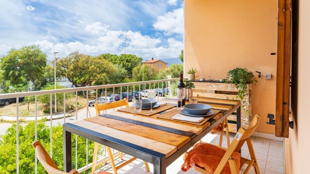 A balcony with a table set for dining, chairs, and a view of the street and trees.
