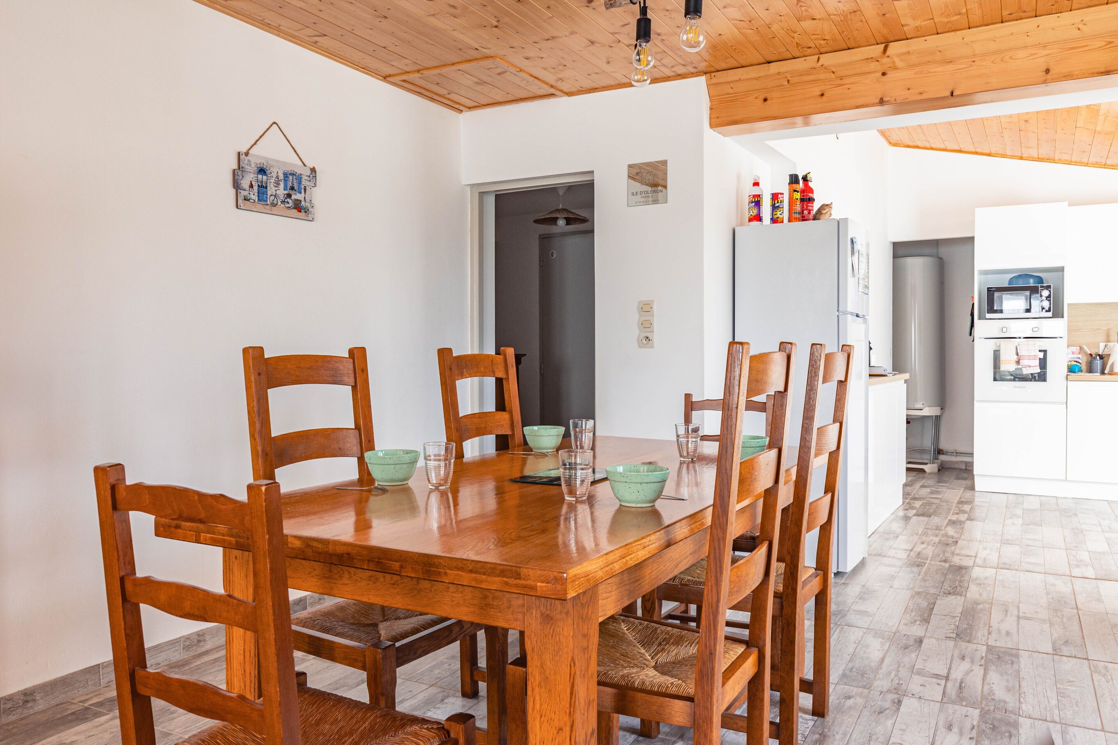 A dining area with a wooden table and chairs, a kitchen in the background, and a hanging light fixture.