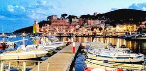 Uma marina com barcos atracados, um prédio à beira-mar e uma colina ao fundo.