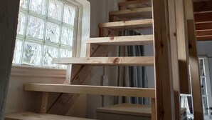 Uma escada de madeira com uma janela à esquerda, teto com vigas aparentes e uma parede em construção.