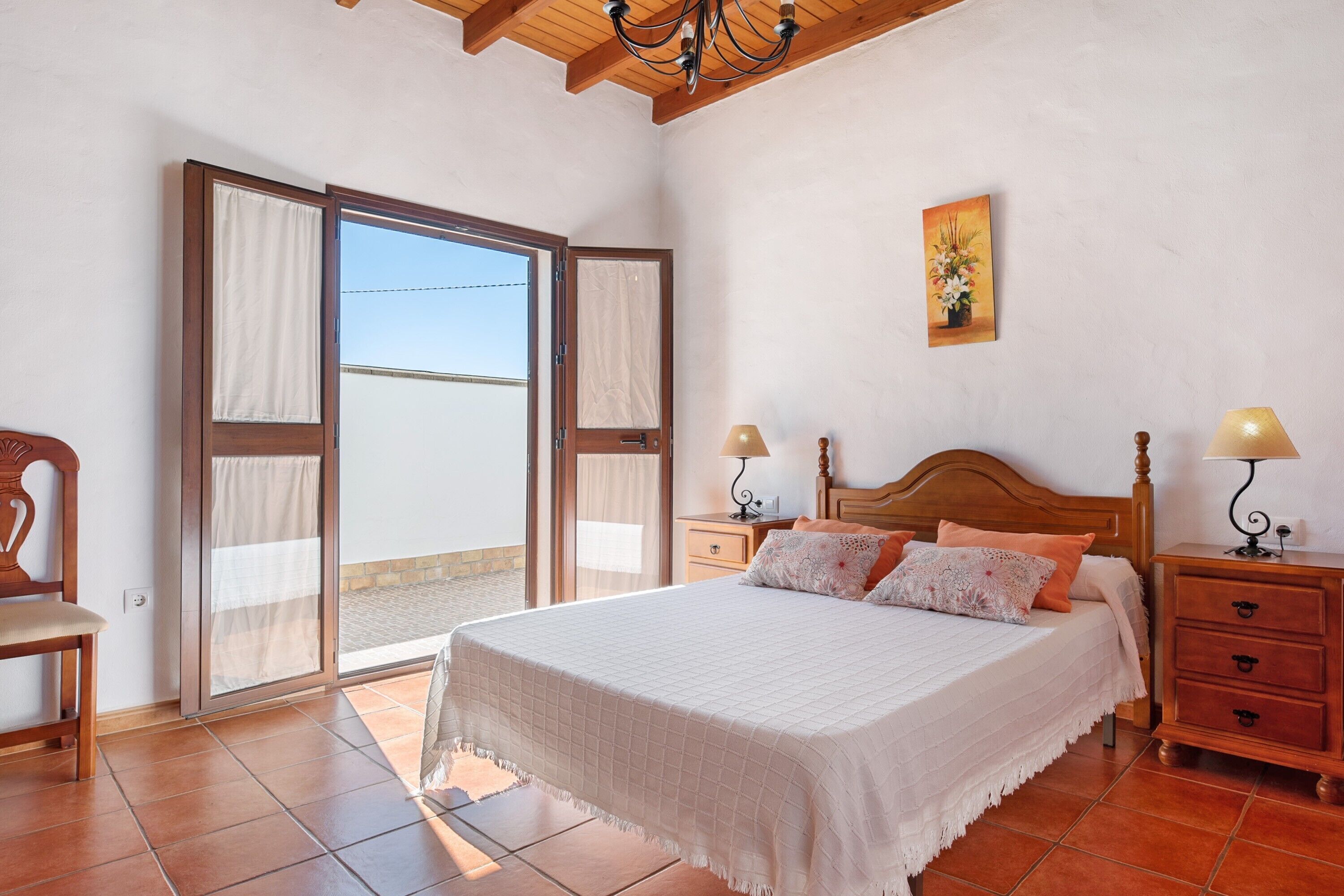 Un dormitorio con una cama, dos mesitas de noche, una silla, un ventanal corredero y un cuadro en la pared.