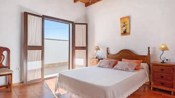 Un dormitorio con una cama, dos mesitas de noche, una silla, un ventanal corredero y un cuadro en la pared.