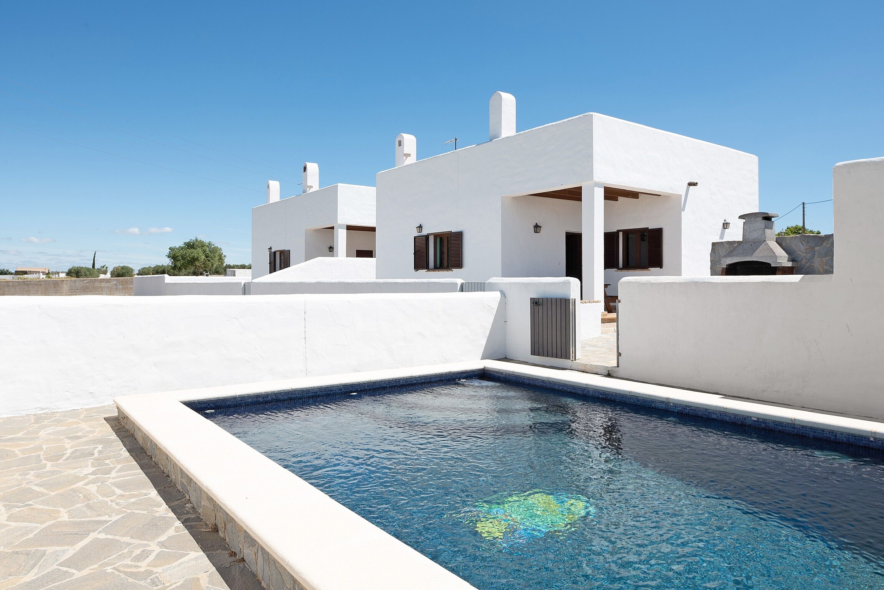 Un edificio moderno y blanco con piscina, suelo de piedra y cielo azul despejado.