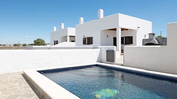 Un edificio moderno y blanco con piscina, suelo de piedra y cielo azul despejado.