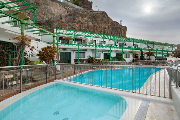 Une piscine agrémentée d’une pergola verte, une falaise rocheuse en arrière-plan et un bâtiment blanc doté de balcons.