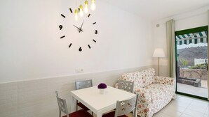 Un coin repas avec une table blanche, des chaises rouges, un canapé à motifs floraux, une horloge murale et une fenêtre donnant sur un paysage.