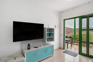 Un salon moderne doté d’un téléviseur à écran plat, de meubles de couleur turquoise, d’un coin repas avec une table et des chaises, et d’une vue sur l’extérieur, visible à travers une porte à cadre vert.