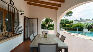 Une terrasse extérieure avec une table à manger, des chaises, une horloge et une piscine en arrière-plan.