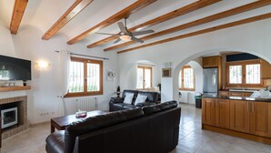 Un vaste espace de vie comprenant un canapé en cuir, des poutres apparentes, un ventilateur de plafond et une cuisine avec des meubles en bois.