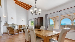 A spacious dining area with a wooden table and chairs, a chandelier, and a view through large windows. - Villa 'Cristina' with Mountain View, Wi-Fi and Air Conditioning (Xàbia)