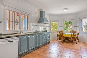 A kitchen with white cabinets, a built-in dishwasher, a blue range hood, a dining table with chairs, and a window with a view of greenery. - Villa 'Cristina' with Mountain View, Wi-Fi and Air Conditioning (Xàbia)
