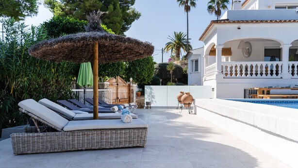 Area piscina con poltrone prendisole, un divano in vimini, un ombrellone di paglia e vista su un edificio bianco con un balcone.