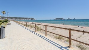 Un sendero pavimentado con baranda de madera que conduce a una playa, cielo azul despejado y formaciones rocosas en la distancia.