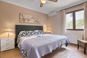 A bedroom with a bed, bedside lamps, a wall-mounted air conditioner, a window with curtains, and a wicker chair. - Villa 'Nichola' with Private Pool, Wi-Fi and Air Conditioning (Xàbia)