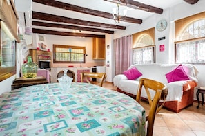 A cozy room with a dining table, a sofa, a wooden chair, a kitchen area, and a window with curtains.