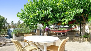 En udendørs patio med et rundt bord og fletstole, omgivet af grønt og en hængekøje.