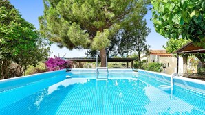 Et klart, blåt swimmingpool med fliseterrasse, omgivet af træer og et overdækket område.