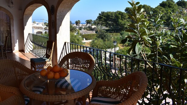 Ein Balkon mit Korbmöbeln, einem Glastisch mit Orangen und Blick auf das Meer und Grünflächen.