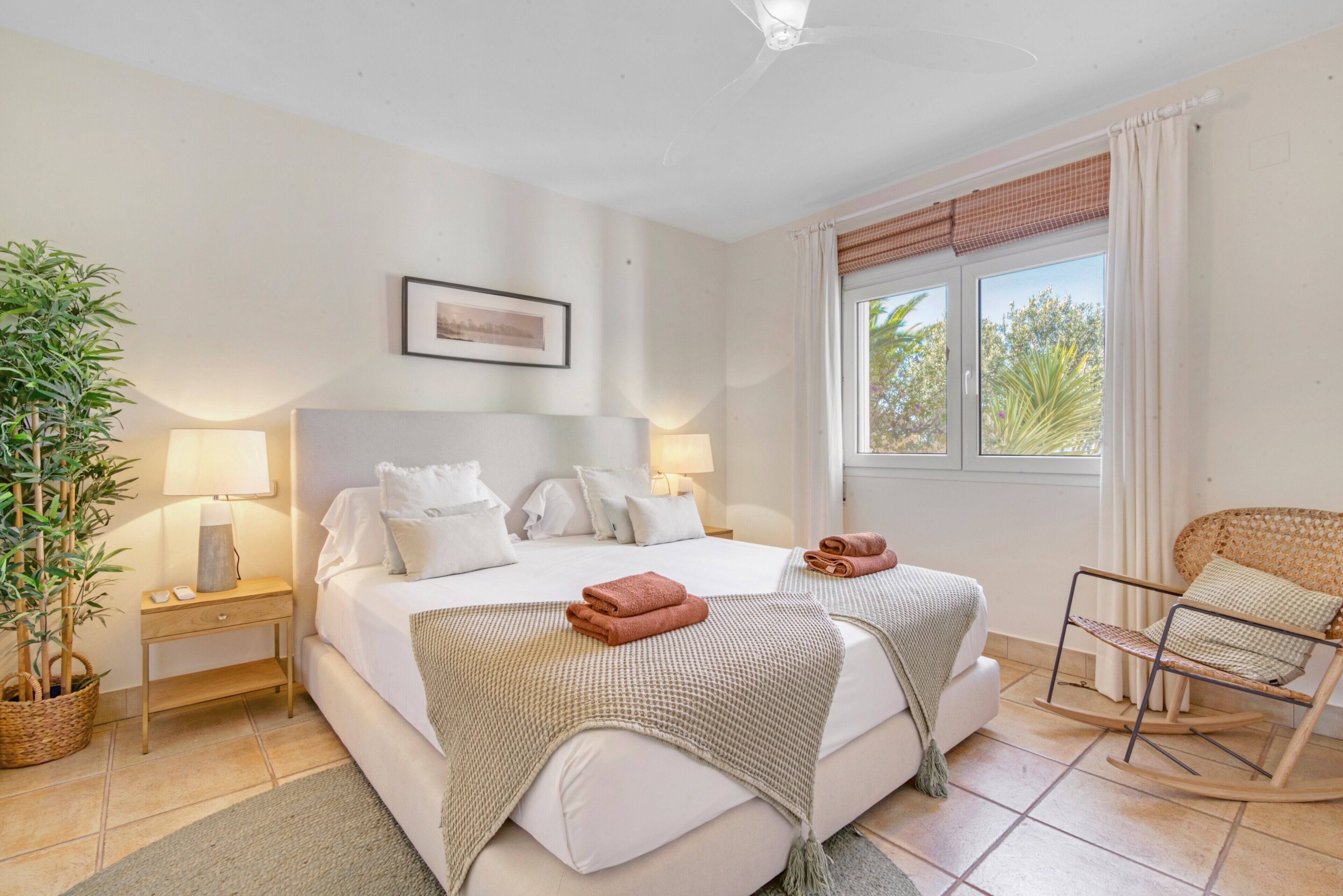 A bedroom with a bed, two bedside tables, a lamp, a rocking chair, a potted plant, and a window with curtains.