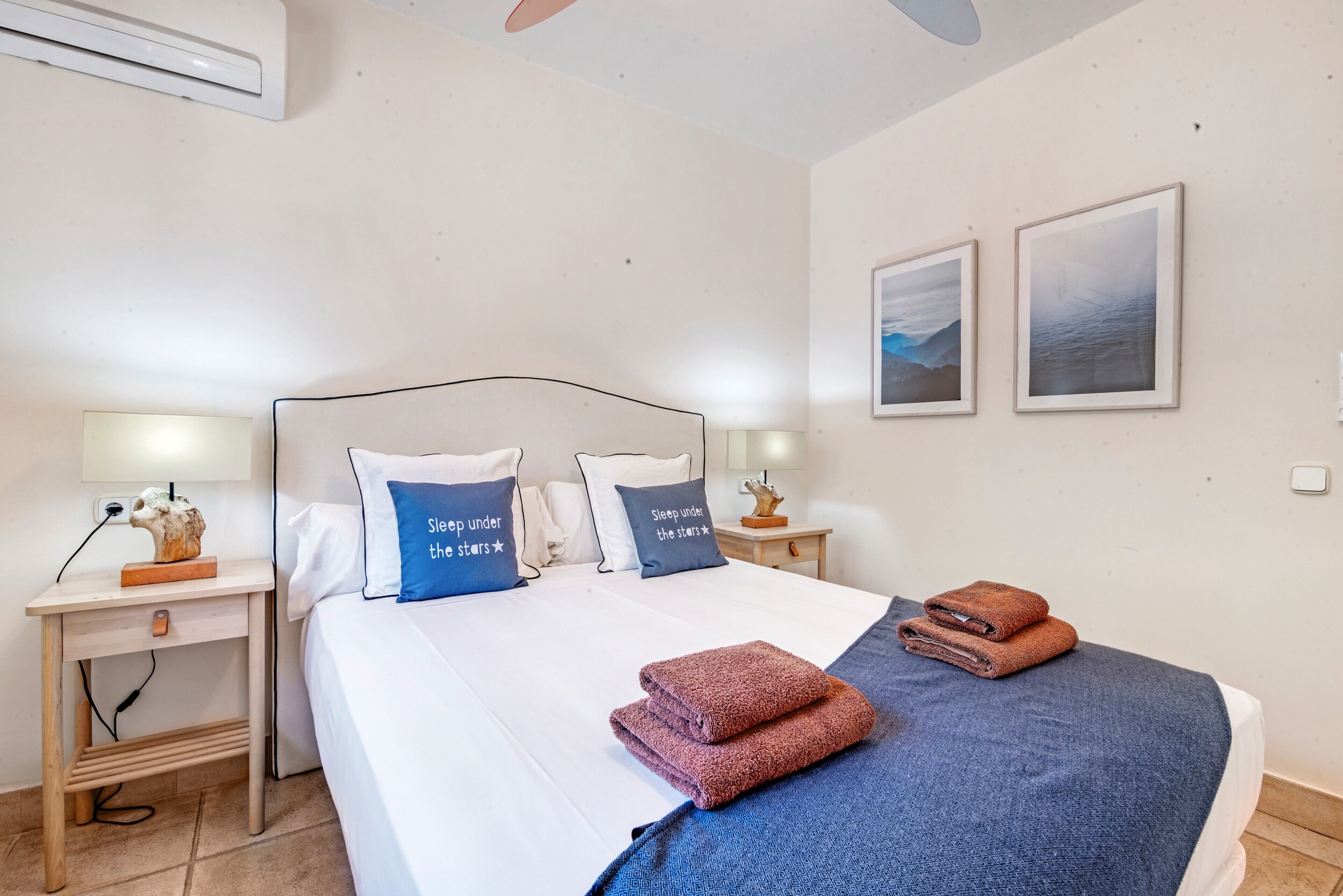 A neatly made bed with pillows, a bedside table with a lamp, and two framed pictures on the wall.
