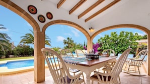A covered outdoor dining area with a pool, white chairs, and a wooden table.