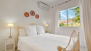 A bedroom with a bed, two bedside tables with lamps, an air conditioning unit, and a window with curtains.