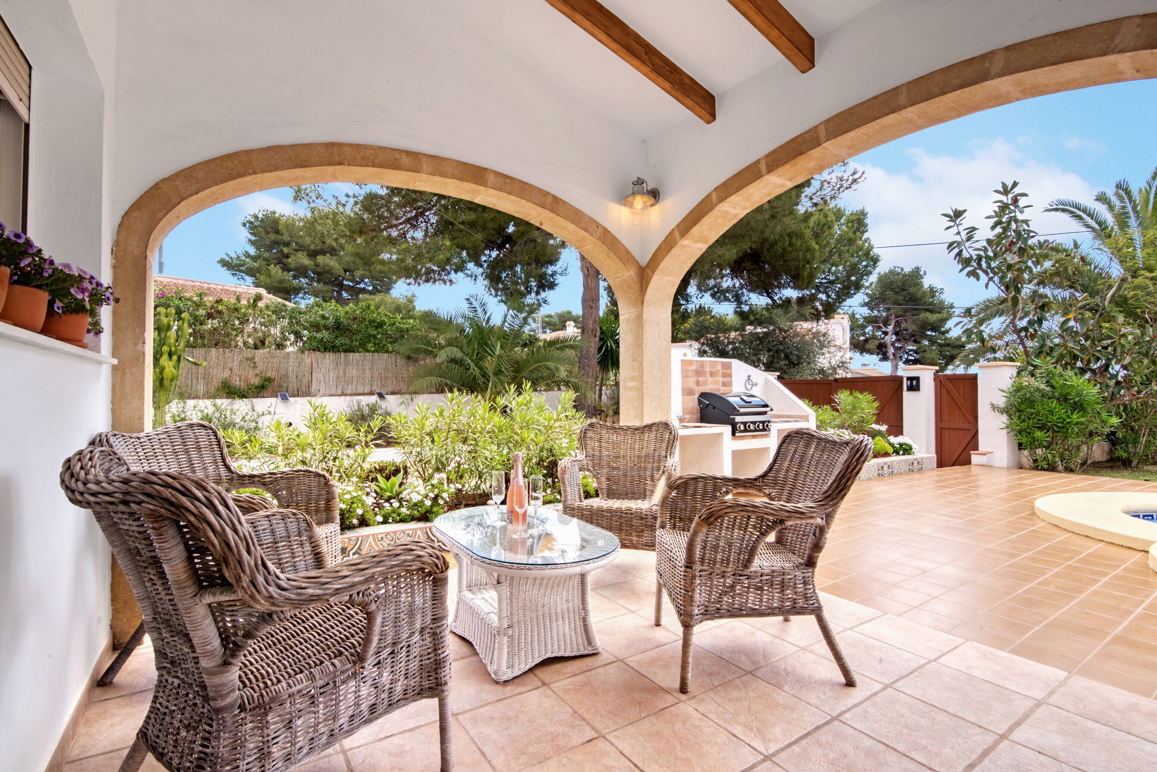 An outdoor patio with wicker furniture, a glass table, and a view of the surrounding garden and trees.