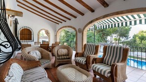 A covered patio area with wicker furniture, wooden beams, and a view of a swimming pool. - Villa 'Rosa' with Private Pool, Shared Terrace and Wi-Fi (Jávea)