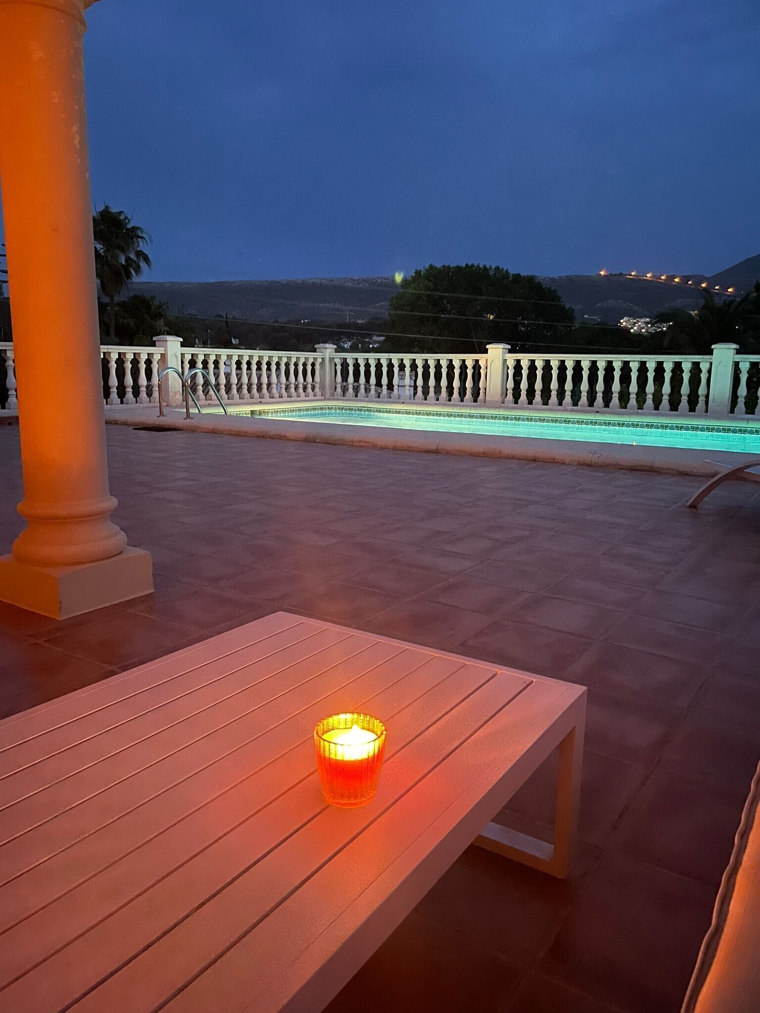 Une terrasse avec un banc et une bougie allumée, d’où l’on peut admirer une piscine et des lumières scintillantes au loin.