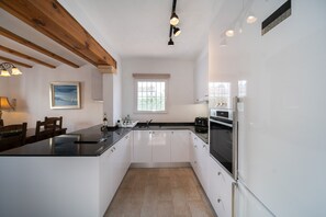 A modern kitchen with white cabinets, a black countertop, wooden beams, a window, and a refrigerator. - Villa 'Sol' with Mountain View, Wi-Fi and Air Conditioning (Alicante)