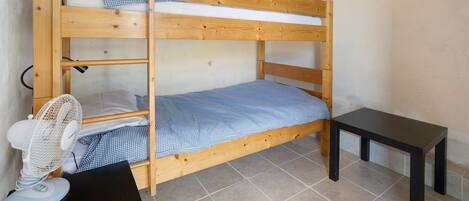 Una cama individual sobre otra, con un ventilador, una mesita y una colcha a cuadros.