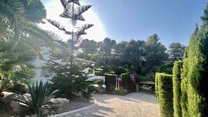 Un chemin de pierres mène à un bâtiment à la façade blanche, entouré de haies soigneusement taillées et de diverses plantes.