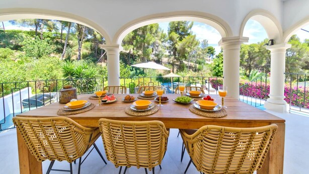 Une table à manger entourée de chaises en osier, mise pour le petit-déjeuner avec des assiettes, des verres et un panier de fruits.