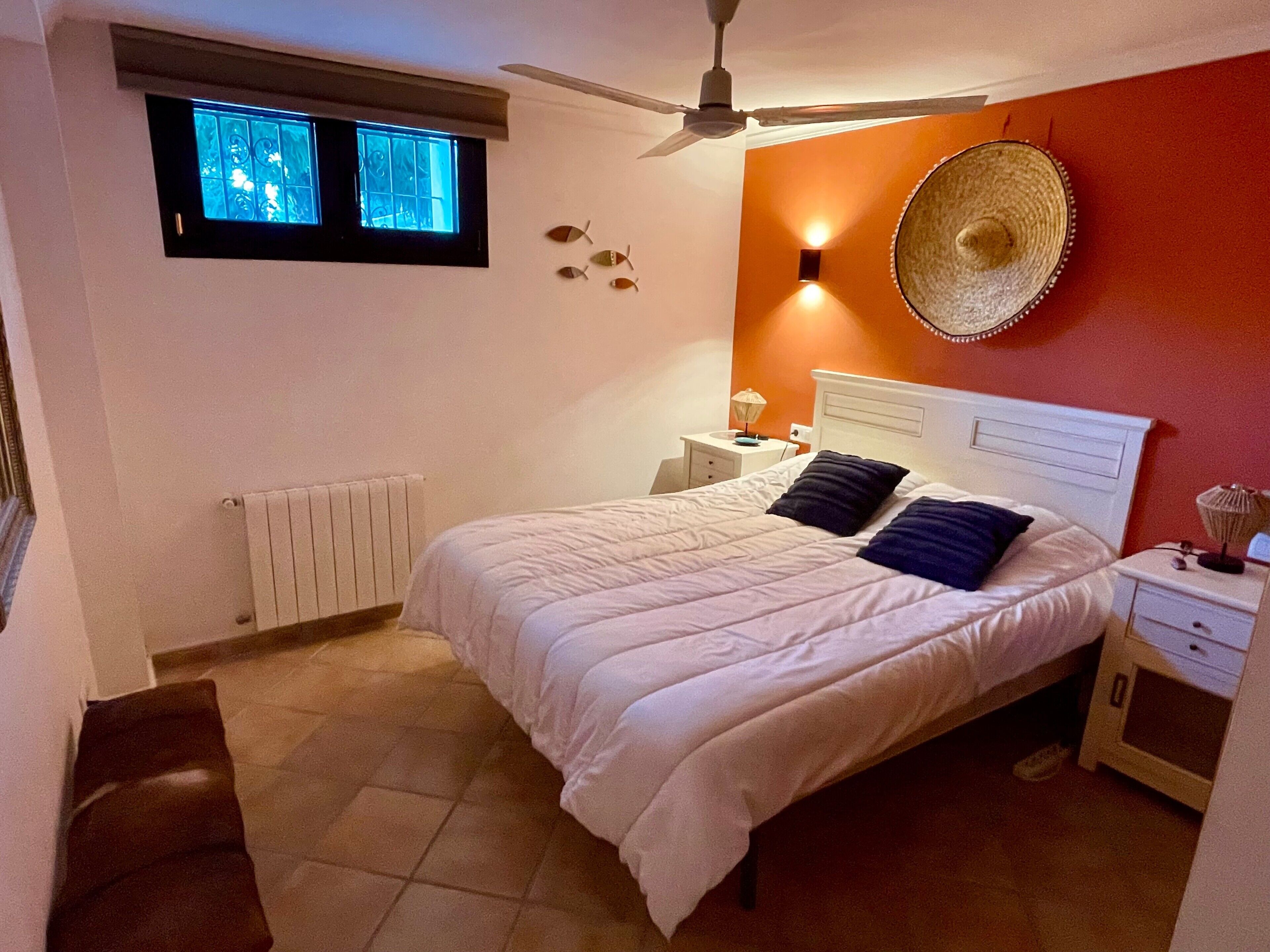 Une chambre à coucher comprenant un lit, un ventilateur de plafond, une fenêtre, un radiateur, une table de chevet et une lampe fixée au mur.