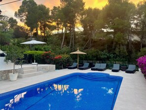 Une piscine avec des chaises longues, une pergola et une vue sur les arbres et le coucher de soleil.