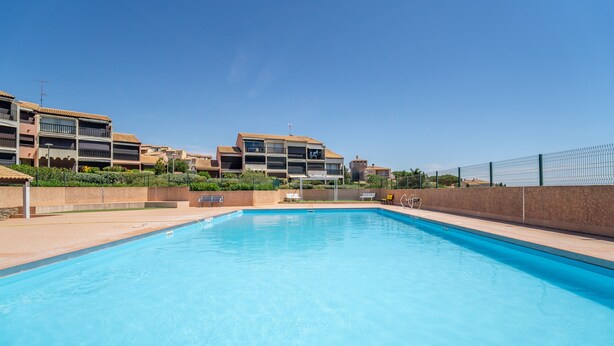 A swimming pool with a clear blue water surface, surrounded by a fence, and adjacent to a building with balconies and a flat roof. - Holiday Home 'Les Eaux Vives 1' with Sea View, Shared Pool and Private Terrace (Fleury)