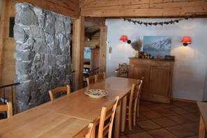 Une table à manger en bois avec des chaises, un mur en pierre, des meubles en bois et deux lampes fixées au mur.