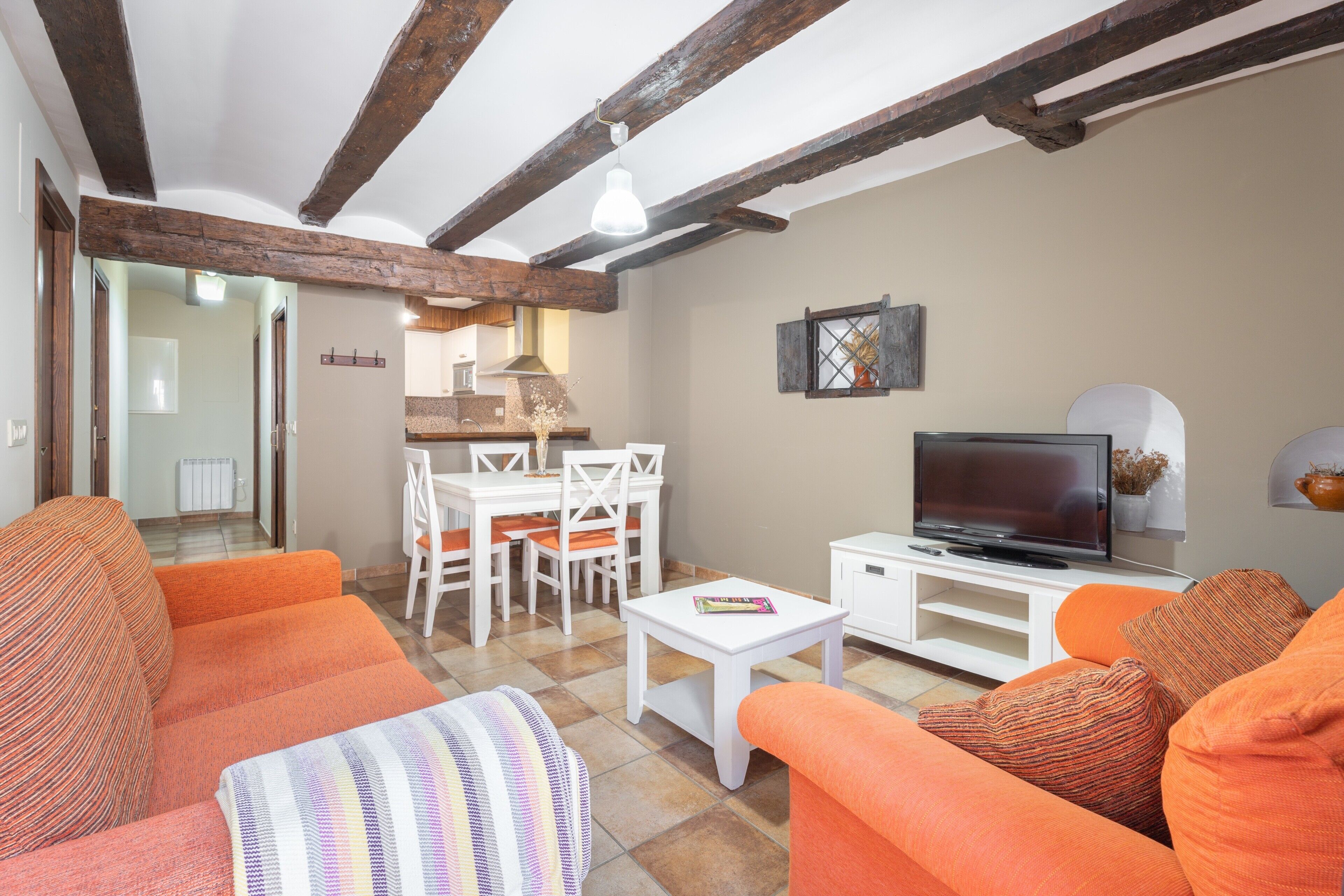 Una sala de estar con sofás naranja, un televisor, un comedor con sillas blancas y vigas de madera a la vista en el techo.