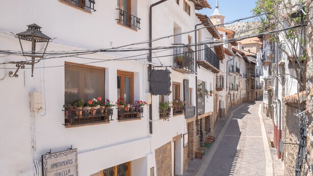 Une rue étroite bordée de bâtiments blancs, de balcons ornés de plantes en pot et de panneaux indiquant « APPARTEMENTS CORELLA » et « BAR-RESTAURANT Corella ».