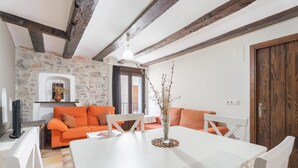 Un espace repas avec une table blanche, un canapé orange, un mur en pierre, des poutres en bois et une porte en bois.