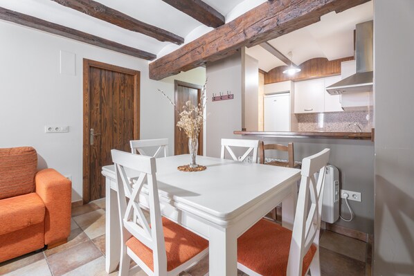 Un comedor con una mesa y sillas blancas, una puerta de madera y una cocina al fondo.