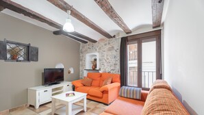 Un salon avec un mur en pierre, un téléviseur posé sur une table blanche, un canapé orange et un balcon avec vue.