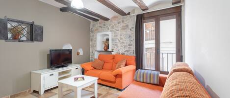 A living room with a stone wall, a television on a white table, an orange sofa, and a balcony with a view.