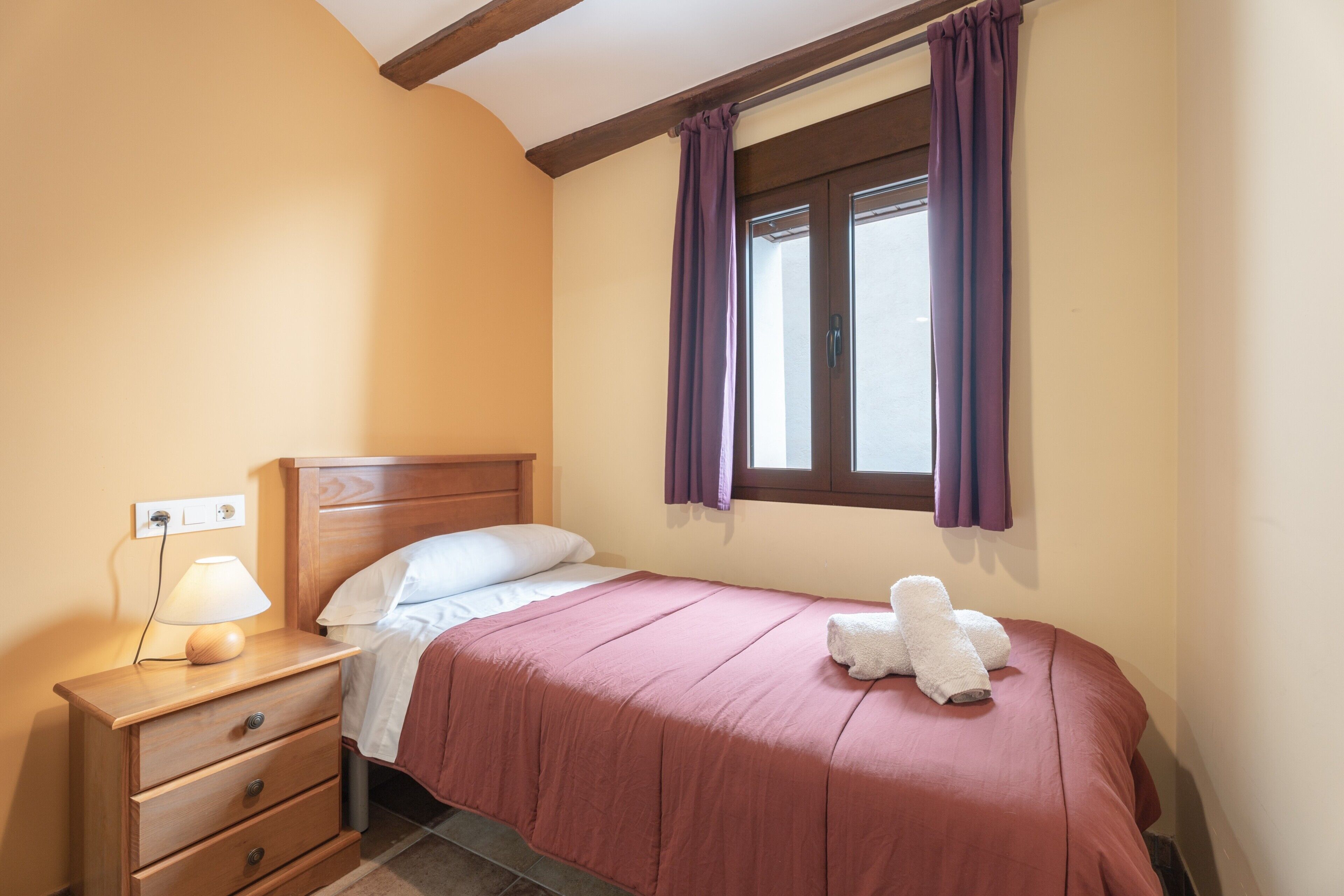 Een slaapkamer met een bed, nachtkastje, lamp en een raam met gordijnen.