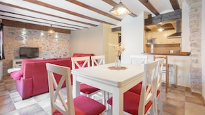 Un coin repas avec une table blanche et des chaises rouges, un mur en pierre et une cuisine en arrière-plan.
