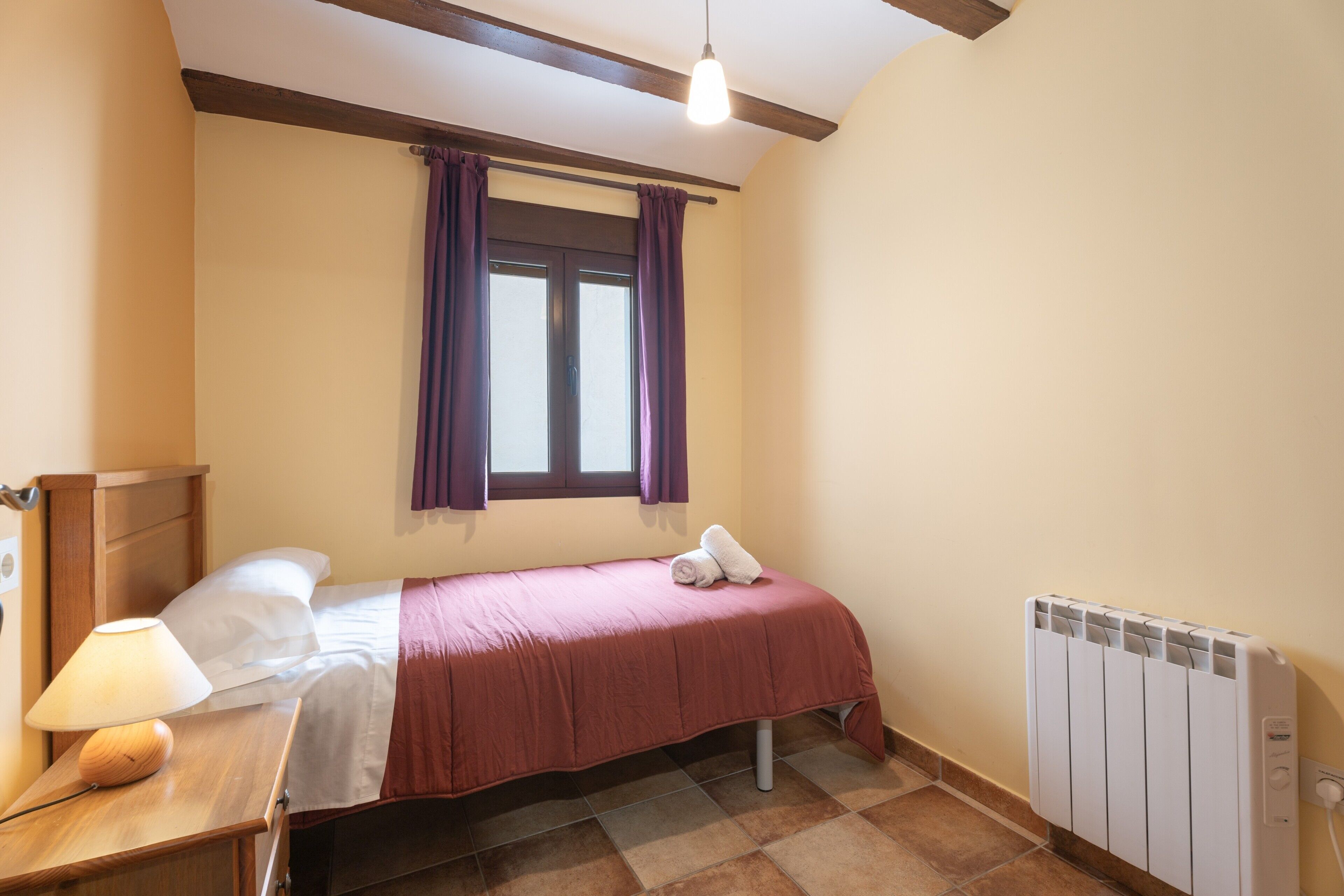 A single bed with a wooden headboard, a bedside table with a lamp, a window with purple curtains, and a radiator.