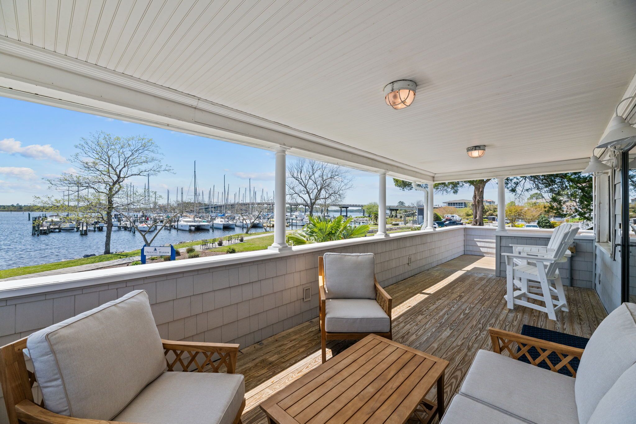 A covered outdoor area with wooden flooring, white cushioned chairs, and a table. A view of the marina with boats and a bridge is visible through the windows.