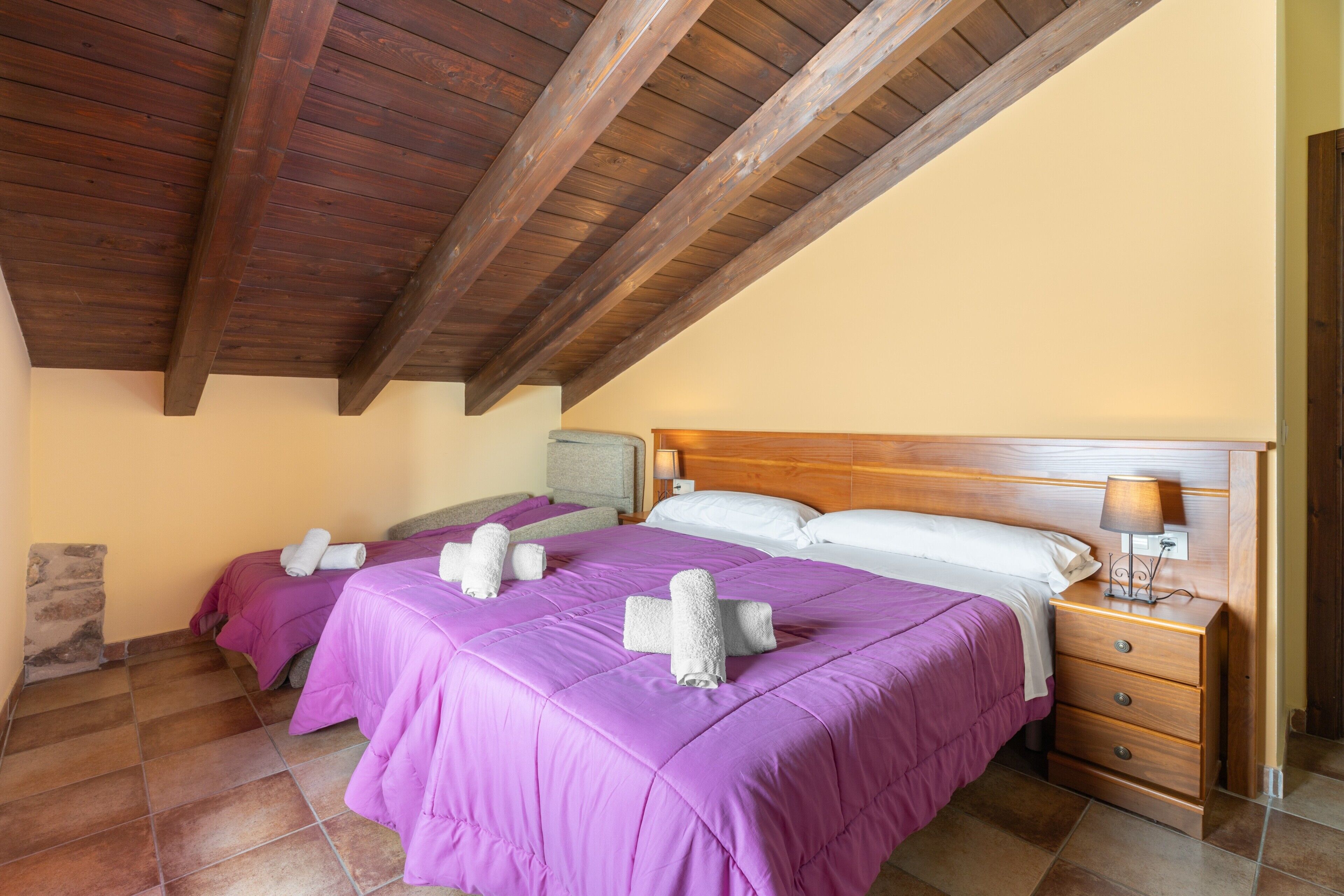 A bedroom with two beds, wooden headboards, a nightstand with a lamp, and a stone wall.