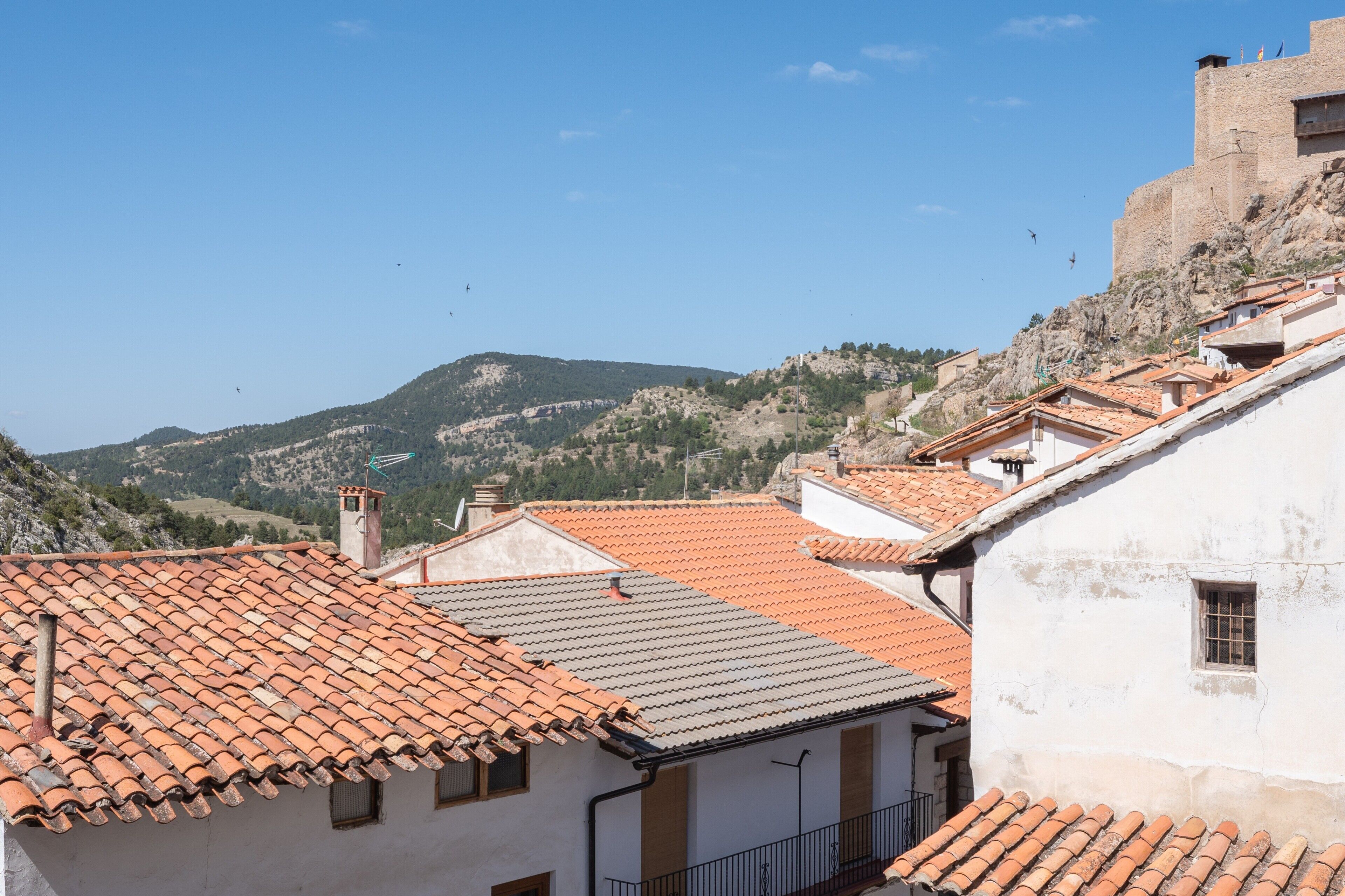 Blick auf terrakottafarbene Dächer, ein Gebäude mit einer Steinmauer und eine gebirgige Landschaft im Hintergrund.